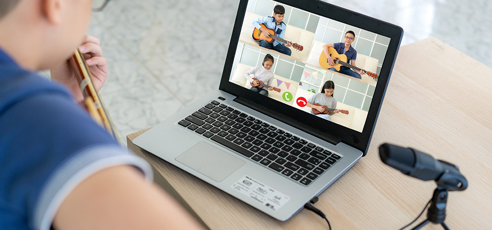 A musician playing the guitar in front of their laptop with four other musicians on the screen also playing instruments. 