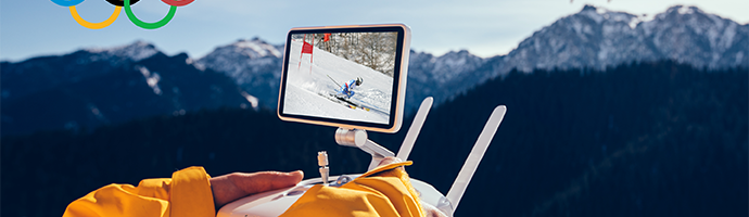 A person in a ski coat operating a drone over snow-covered mountains with an Olympic logo in the top-left corner.