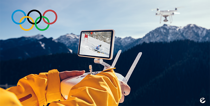 A person in a ski coat operating a drone over snow-covered mountains with an Olympic logo in the top-left corner.